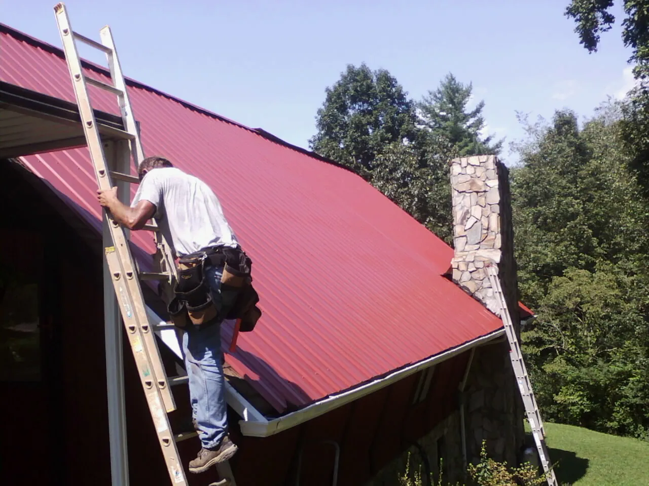 Roofer on a ladder beside a red metal roof and stone chimney — Soffit & Fascia Repair by Roofing Brothers in Warner Robins